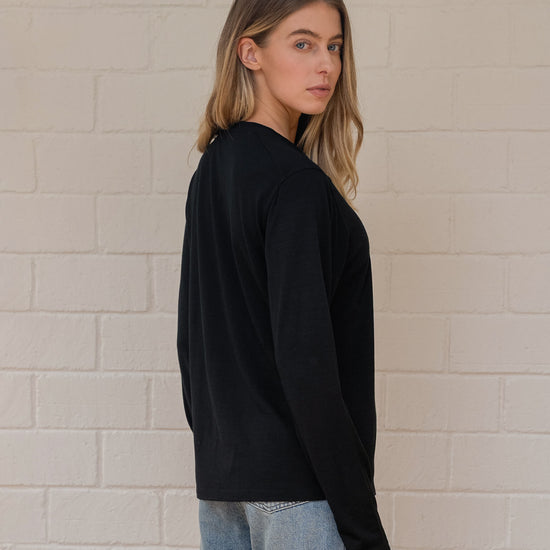 Woman wearing a black long-sleeve top and light blue jeans against a beige brick wall.