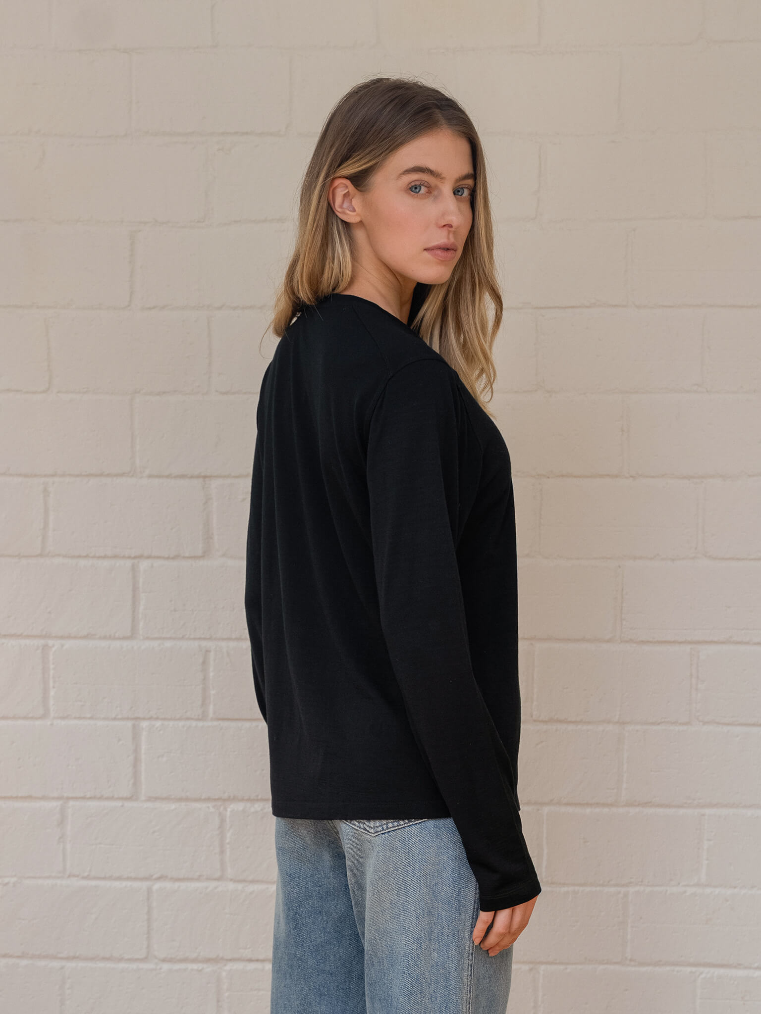 Woman wearing a black long-sleeve top and light blue jeans against a beige brick wall.
