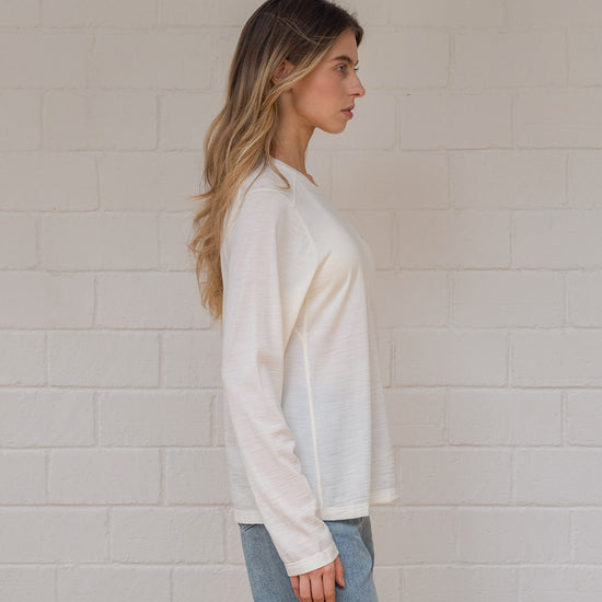 Woman wearing off white long-sleeve shirt and blue jeans against a light brick wall.