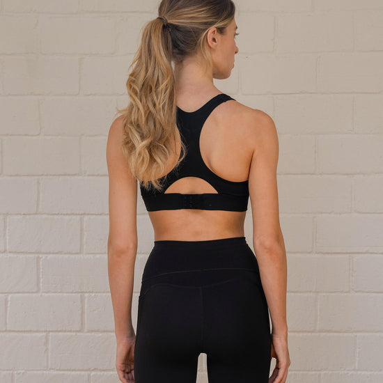 Woman wearing a black racer-back sports bra with adjustable back closure and matching leggings facing the wall.