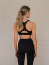 Woman wearing a black racer-back sports bra with adjustable back closure and matching leggings facing the wall.