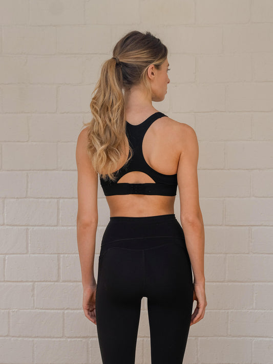 Woman wearing a black racer-back sports bra with adjustable back closure and matching leggings facing the wall.