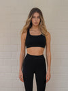 Woman wearing a black sports bra and matching leggings against a white wall.