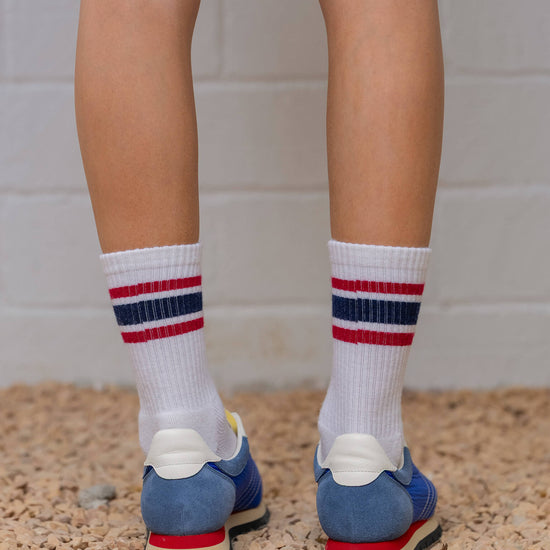 Person wearing white socks with red and blue stripes and blue shoes on a stone surface.