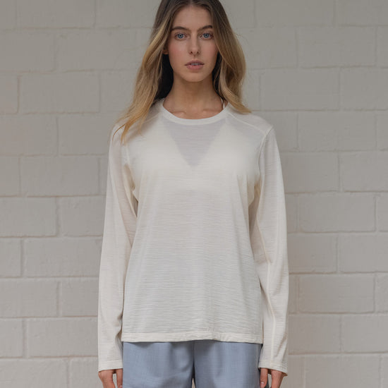 Woman wearing off white long-sleeve shirt and light blue shorts against a gray brick wall.