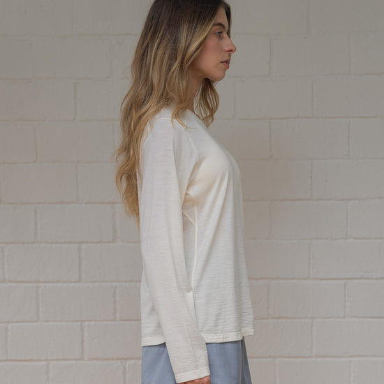 Woman wearing off white long-sleeve top and light blue skirt against a white brick wall.