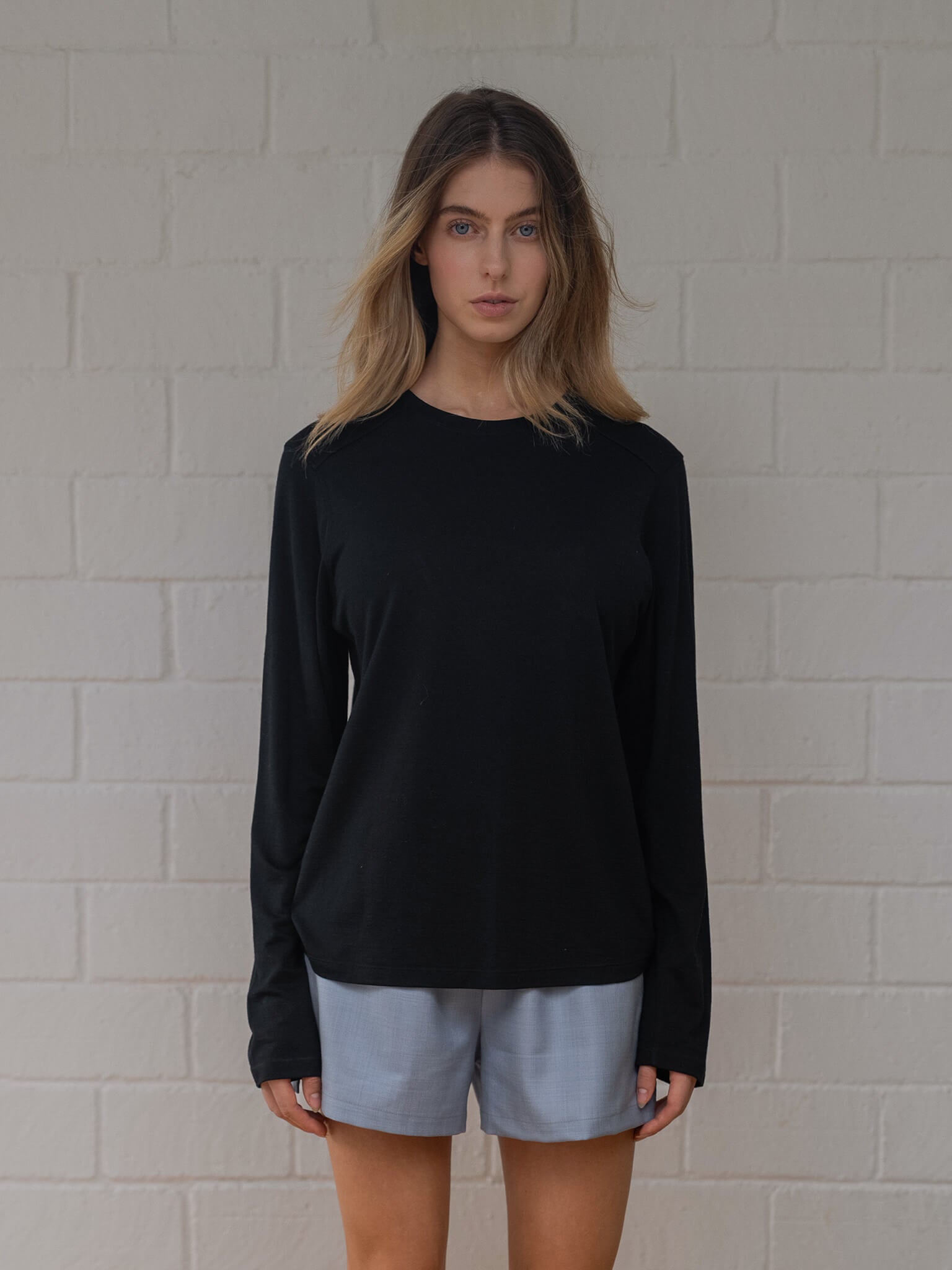 Person wearing a black long-sleeve shirt and light blue shorts against a white brick wall.