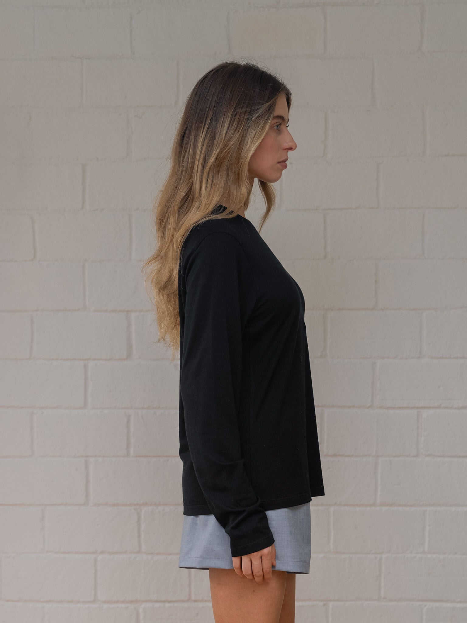 Woman wearing a black long-sleeve top and light blue shorts against a white brick wall.
