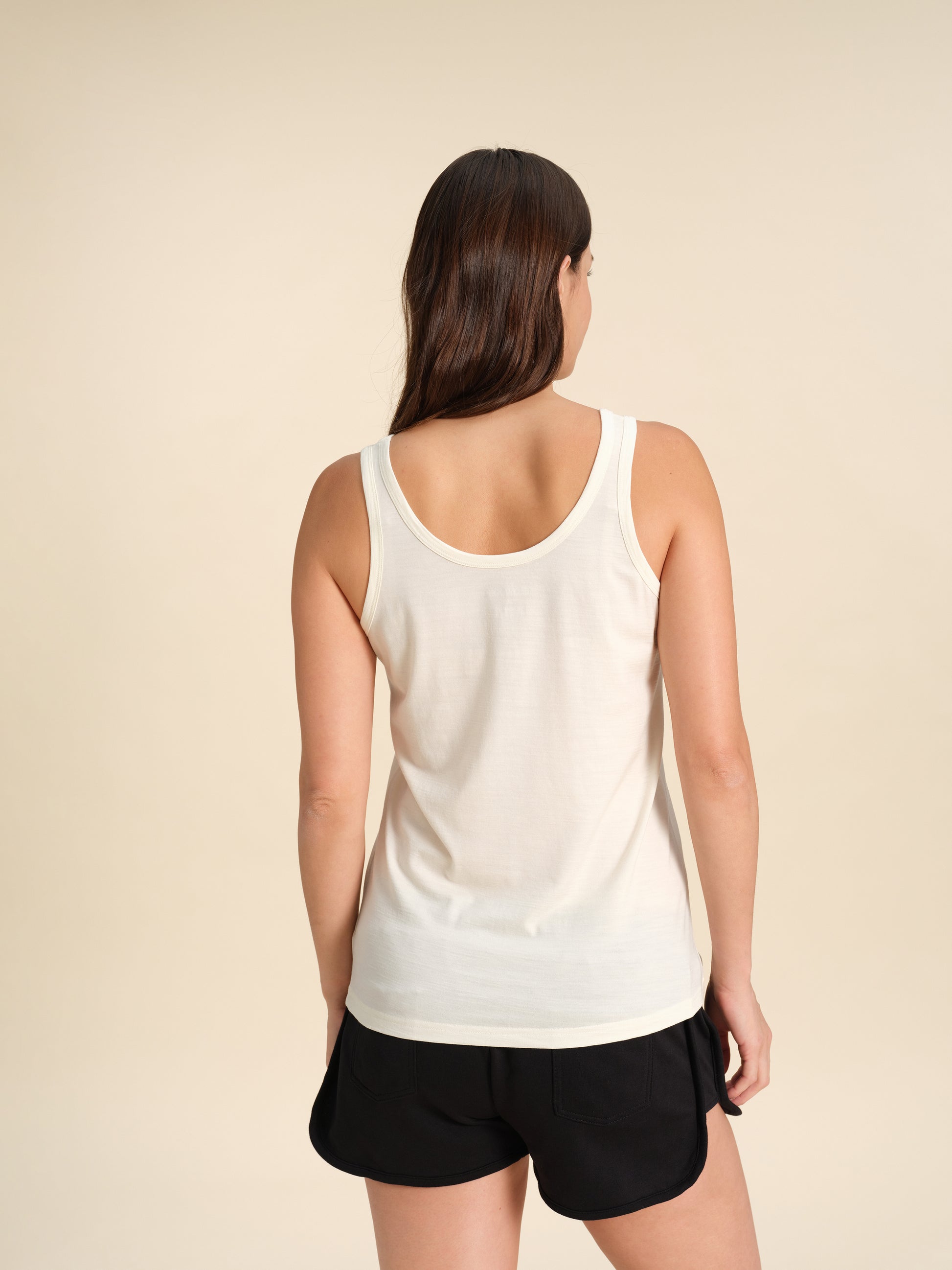 Back view of a woman wearing a white Merino wool scoop tanks against a sand background.